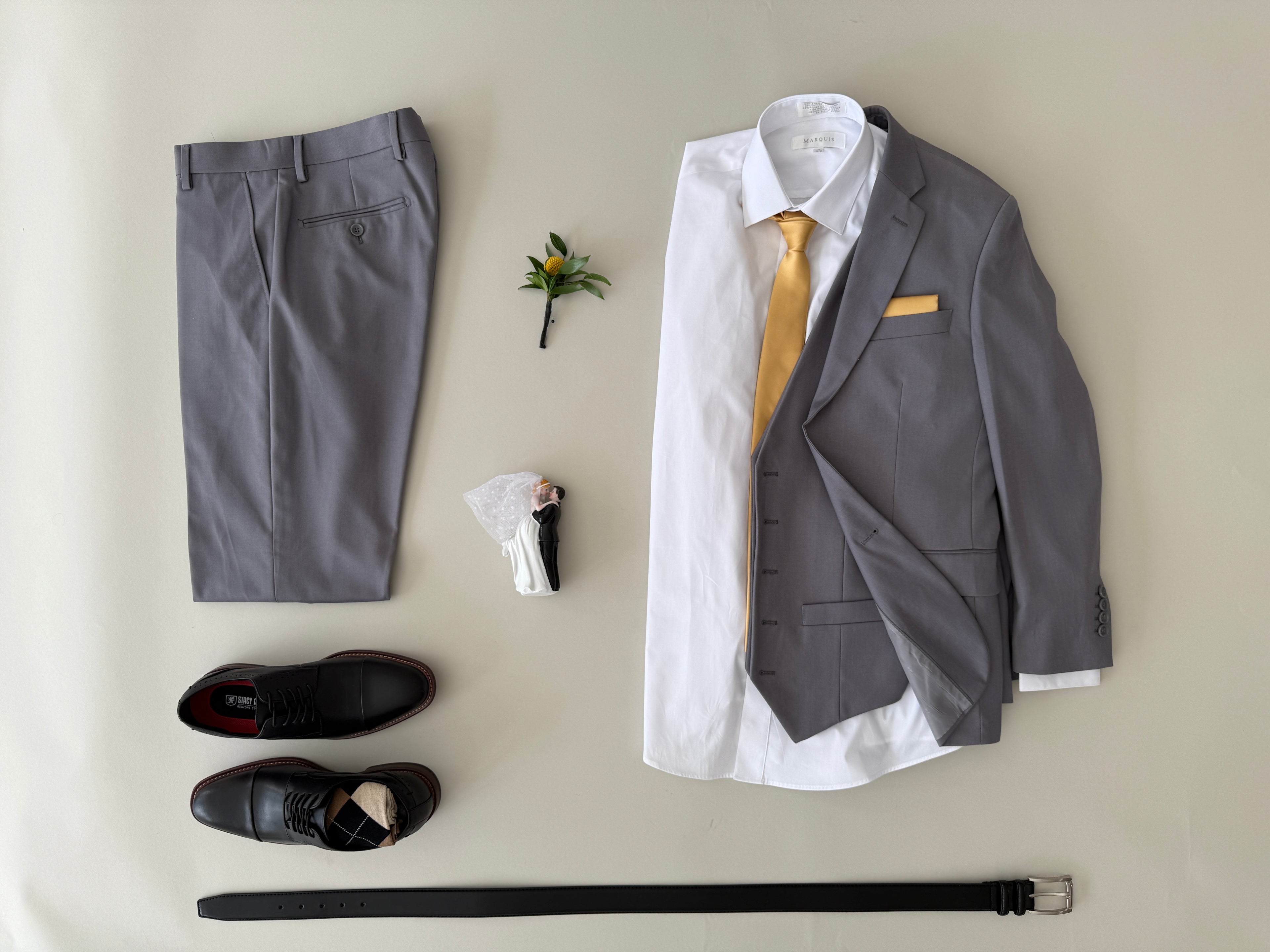 Studio shot light grey vested suit, white dress shirt, gold tie and pocket square, black dress shoes and black belt.