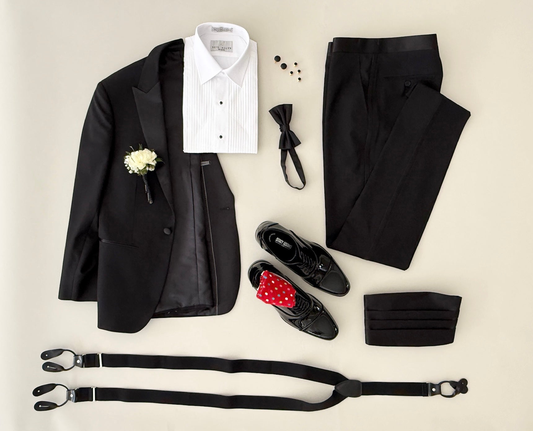 Studio image of a black peak lapel tuxedo, pleated laydown formal shirt, black bowtie and cummerbund, gold cufflinks and studs, patent leather formal shoes and black leather trim suspenders.